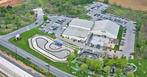 Aerial view of a go-kart track, mini-golf, and a large parking lot near a building surrounded by greenery.