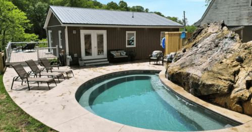 A backyard with a curved pool, lounge chairs, a rock feature, and a small cabin in a green, wooded area.