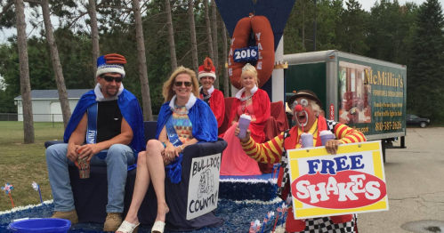 A festive parade float with people in costumes, including a clown holding a "Free Shakes" sign, celebrating a local event.