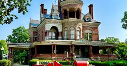 A large, ornate Victorian mansion with a red vintage fire truck parked in front, surrounded by lush greenery.