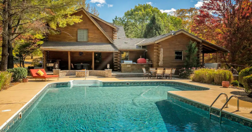 A log cabin home with a pool in the foreground, surrounded by trees and autumn foliage.