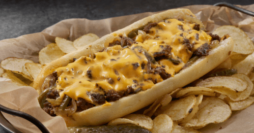 A loaded cheesesteak sandwich with melted cheese, served with crispy potato chips on a parchment-lined tray.
