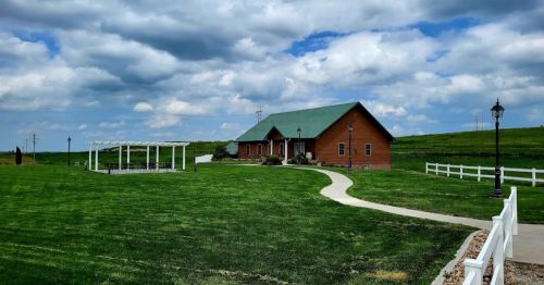 A wooden house with a green roof sits on a grassy lawn under a cloudy sky, with a pathway and lampposts nearby.