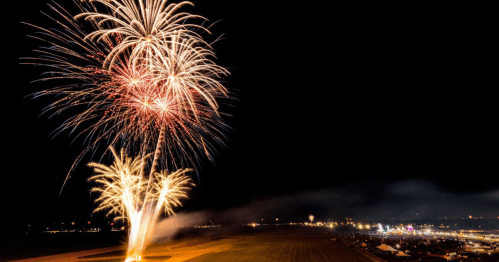 Colorful fireworks burst in the night sky over a field, illuminating a distant festival with vibrant lights.