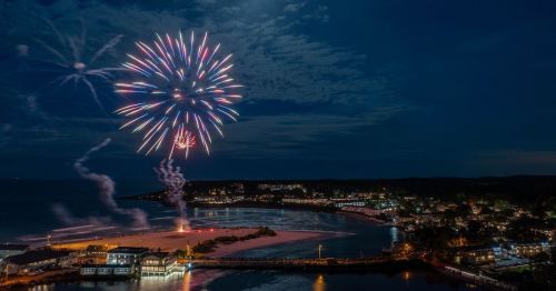 Colorful fireworks illuminate the night sky over a coastal town, reflecting on the water and highlighting the shoreline.