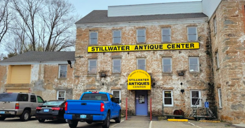 Exterior of the Stillwater Antique Center, featuring a weathered building with a bright yellow sign and parked vehicles.