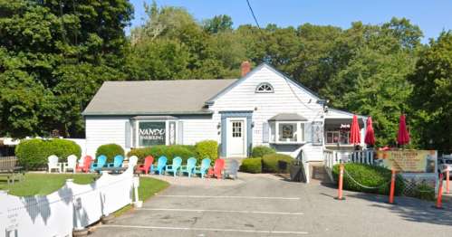 A charming white building with a gray roof, surrounded by colorful chairs and greenery, featuring outdoor seating.