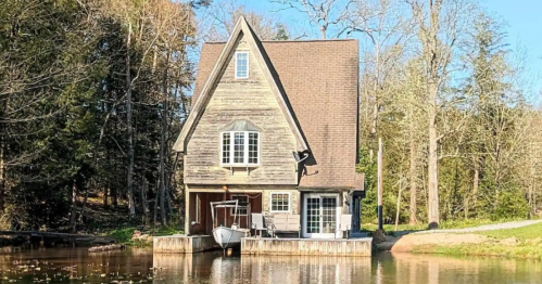 A charming wooden house by a calm lake, surrounded by trees and reflecting in the water under a clear blue sky.