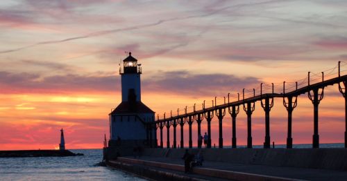 A lighthouse stands by the water at sunset, with vibrant colors in the sky and a pier extending into the lake.