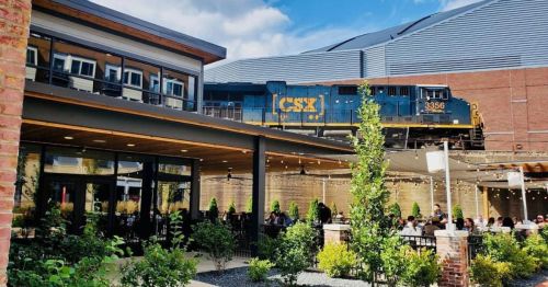 A CSX train passes above a restaurant patio filled with diners, surrounded by greenery and modern architecture.