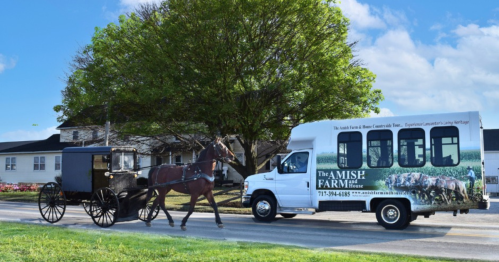A horse-drawn buggy passes a van advertising an Amish farm, with a rural landscape and blue sky in the background.