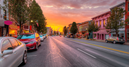 A vibrant sunset casts orange hues over a charming street lined with parked cars and colorful storefronts.