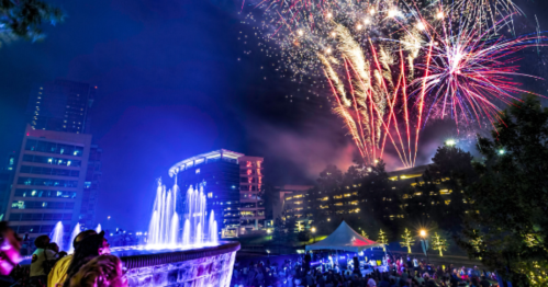 Colorful fireworks illuminate the night sky above a fountain and crowd at a festive outdoor event.