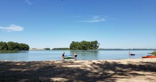 Two people near a calm lake with trees in the background and boats on the water under a clear blue sky.