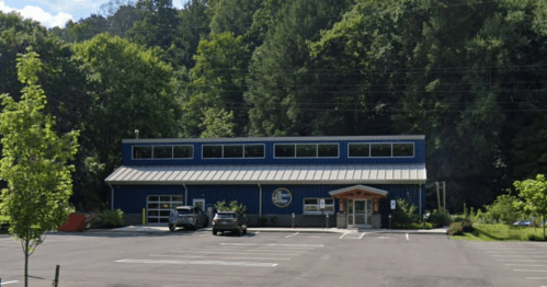 A blue building with large windows, surrounded by trees, and a parking lot with a few cars.