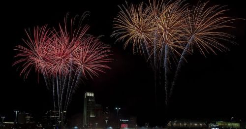 Colorful fireworks burst in the night sky over a city skyline, illuminating the darkness with vibrant reds and golds.