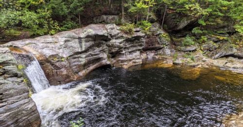 A serene waterfall cascades into a dark pool, surrounded by rocky cliffs and lush green foliage.