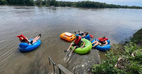 Four people relax on colorful inner tubes in a river, enjoying a sunny day with trees lining the shore.