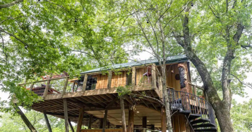 A wooden treehouse elevated among green trees, featuring a spiral staircase and a cozy balcony.