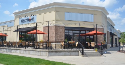 Exterior of a restaurant with a patio, featuring red umbrellas and a modern design, surrounded by green grass.
