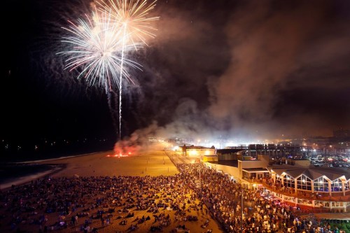 Fireworks illuminate the night sky over a crowded beach, with people gathered along the shore and buildings in the background.