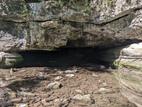 A rocky cave entrance with a dry, cracked ground and sparse vegetation surrounding it.