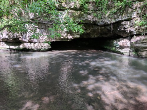 A serene river flows beneath a rocky overhang, surrounded by lush greenery and dappled sunlight.