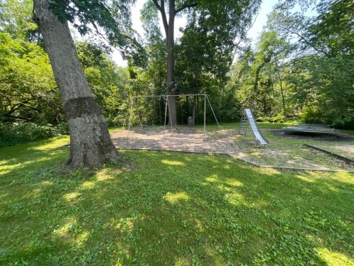 A grassy area with a swing set and slide surrounded by trees in a sunny park.