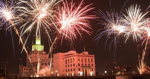 Colorful fireworks illuminate a historic building at night, with a green-lit tower and a festive atmosphere.