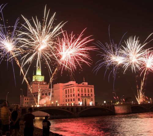 Colorful fireworks illuminate a city skyline at night, reflecting on the water below, with spectators watching from the shore.