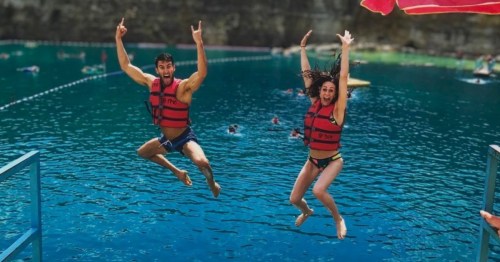 A man and woman in red life jackets excitedly jump mid-air into deep blue water at an outdoor quarry park.