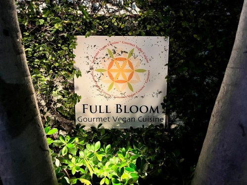 Sign for "Full Bloom Gourmet Vegan Cuisine" surrounded by greenery, featuring a colorful sun design.