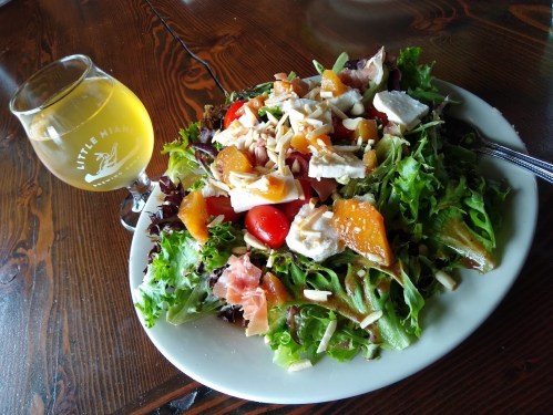 A vibrant salad with greens, cherry tomatoes, chicken, and nuts, served with a glass of light beer on a wooden table.