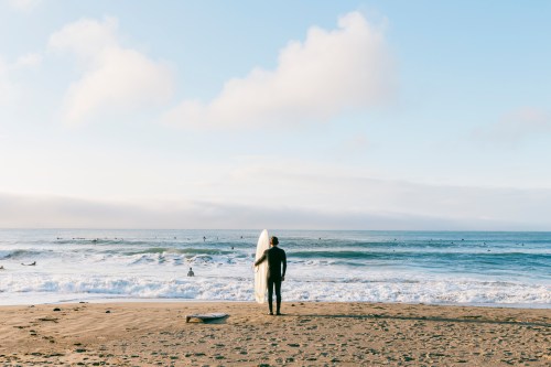 A surfer in a wetsuit walks along the beach holding a surfboard, with waves and other surfers in the background.