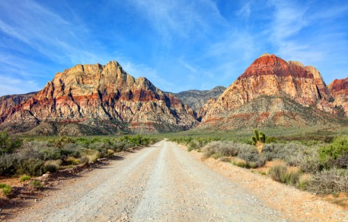 A gravel road leads through a desert landscape with colorful mountains under a blue sky.