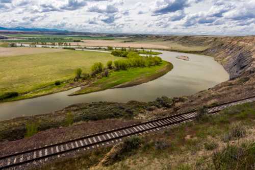 A winding river flows through a green landscape, with rolling hills and a railway track in the foreground under a cloudy sky.