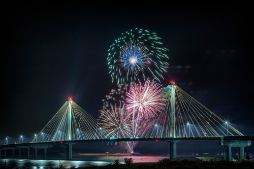 Colorful fireworks illuminate the night sky above a lit bridge, reflecting on the water below.