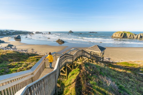 A person in a yellow jacket walks down wooden stairs towards a sandy beach with rocky formations and gentle waves.