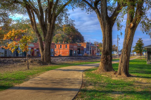 A scenic view of a tree-lined path leading to a quaint town with brick buildings and a railway track.