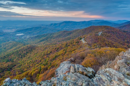 A panoramic view of rolling hills covered in autumn foliage under a colorful sky at sunset.