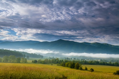 A serene landscape featuring rolling mountains, a misty valley, and a cloudy sky over a lush green field.