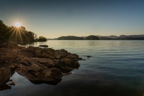 Sunrise over a calm lake, with rocky shorelines and distant hills silhouetted against the morning light.