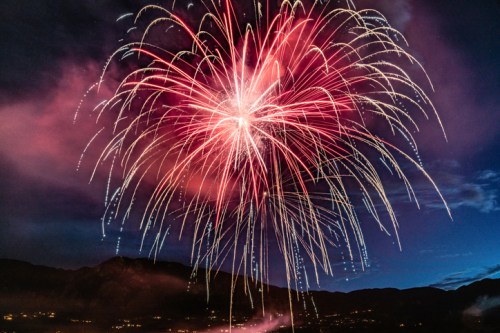 Colorful fireworks burst in the night sky, illuminating clouds and mountains in the background.