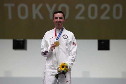 Athlete in a white tracksuit holding a gold medal and flowers, standing on a podium with "Tokyo 2020" in the background.