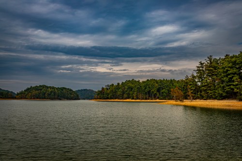 A serene lake surrounded by lush green trees and distant hills under a cloudy sky.