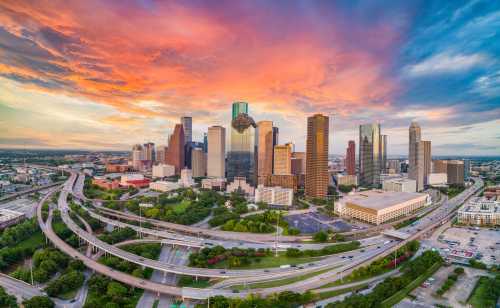 Aerial view of Houston's skyline at sunset, featuring skyscrapers and vibrant clouds over highways and greenery.