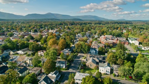 Aerial view of a small town surrounded by mountains, featuring colorful trees and a mix of residential and commercial buildings.