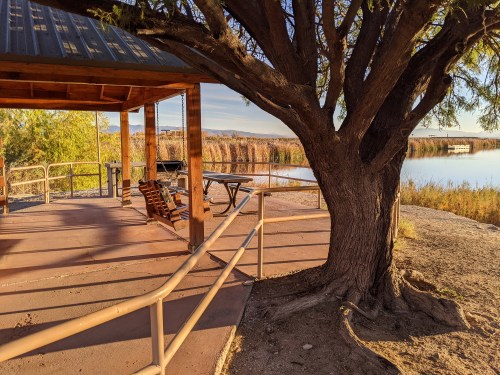 A wooden gazebo by a calm waterway, surrounded by trees and tall grasses, with a bench and picnic table.