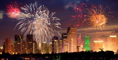 Colorful fireworks illuminate a city skyline at night, with buildings reflecting vibrant lights against a dark sky.