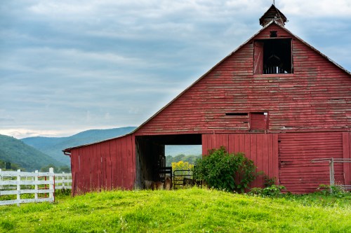A red barn with a sloped roof, surrounded by green grass and hills under a cloudy sky.